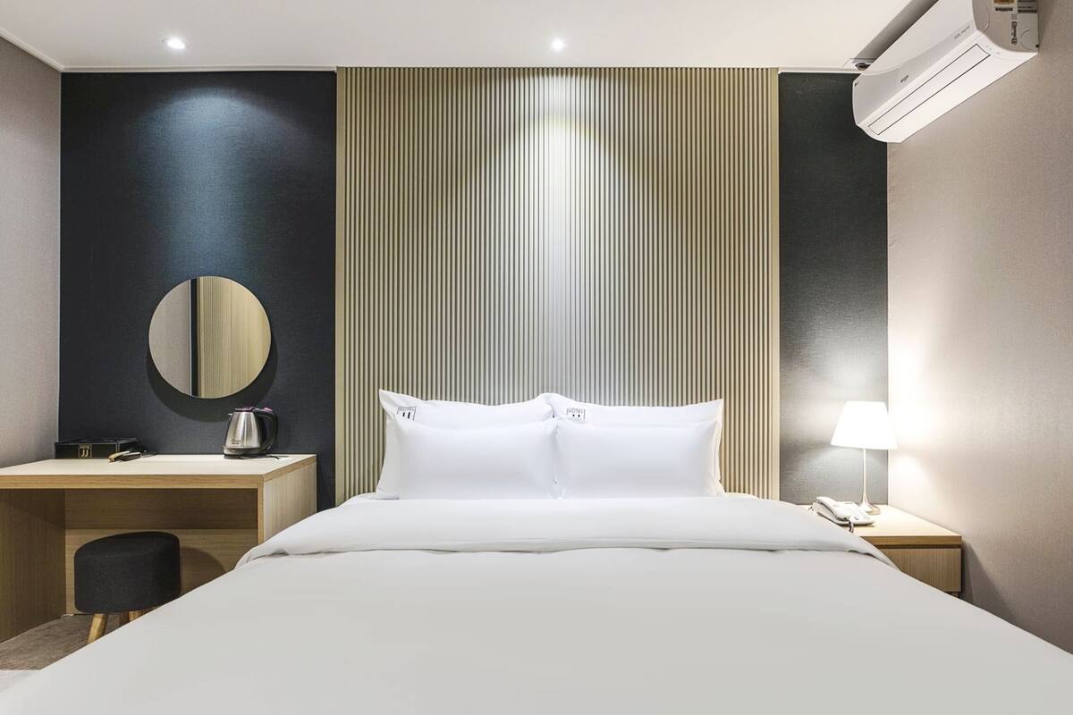 Une chambre d’hôtel comprenant un lit, un bureau, une chaise, un miroir rond, une lampe de chevet et un appareil de climatisation.