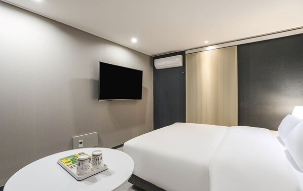 A hotel room with a bed, a flat-screen TV mounted on the wall, a round table with a tray and two mugs, and a window with curtains.