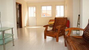 Amplia sala de estar con un sofá y una silla de madera, además de una mesa de centro de vidrio. Tres ventanas proporcionan luz natural.