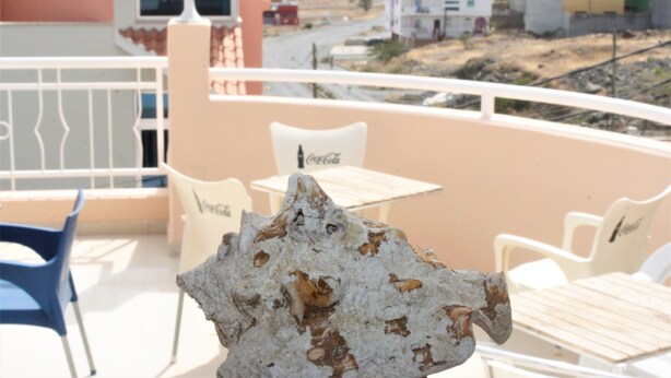 Un balcón con una mesa y sillas blancas, una roca decorativa y vista de edificios al fondo.