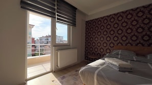 A bedroom with a bed, a radiator, a patterned wall, and a view of buildings through a window with blinds. - Edirne House (Edirne)
