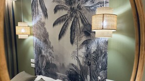 Un dormitorio con una cama, dos almohadas, una mesita de noche, un espejo y un papel tapiz con motivos tropicales.