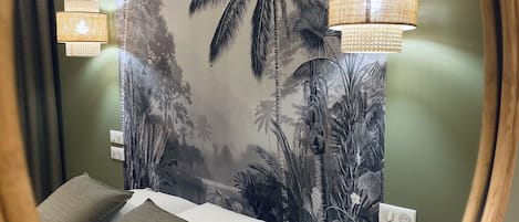 Een slaapkamer met een bed, twee kussens, een nachtkastje, een spiegel en een muur met tropische behang.