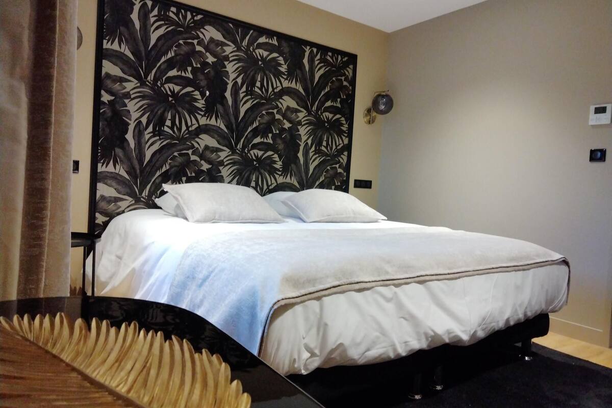 Een opgemaakt bed met witte lakens, een hoofdbord met een tropisch bladmotief en een donkere vloer.