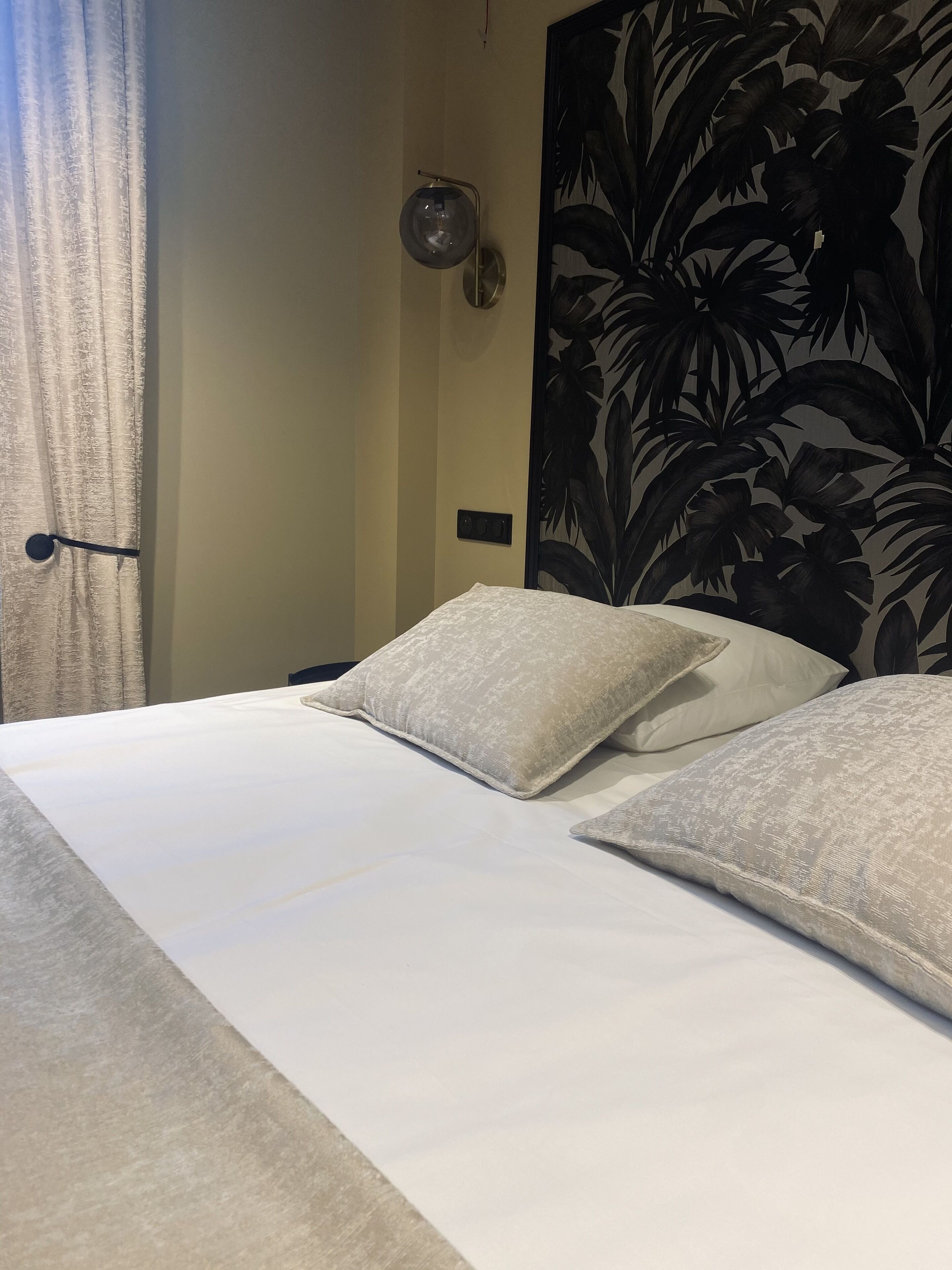 Una cama bien hecha con sábanas blancas y dos almohadas, una lámpara de pared y un papel tapiz con un gran estampado floral.