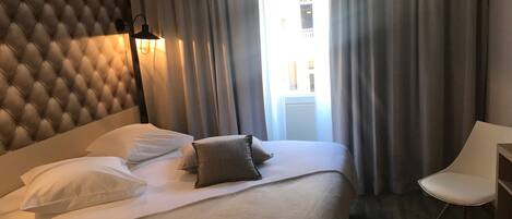 Una habitación de hotel con una cabecera capitonada, una cama con ropa de cama blanca y gris, una lámpara de noche y una silla.