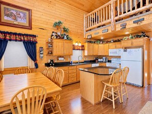 Interior de una cabaña de madera con comedor, cocina y habitación en un entrepiso.