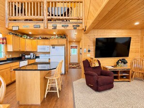 Interior de una cabaña con cocina, comedor y sala de estar con televisor.