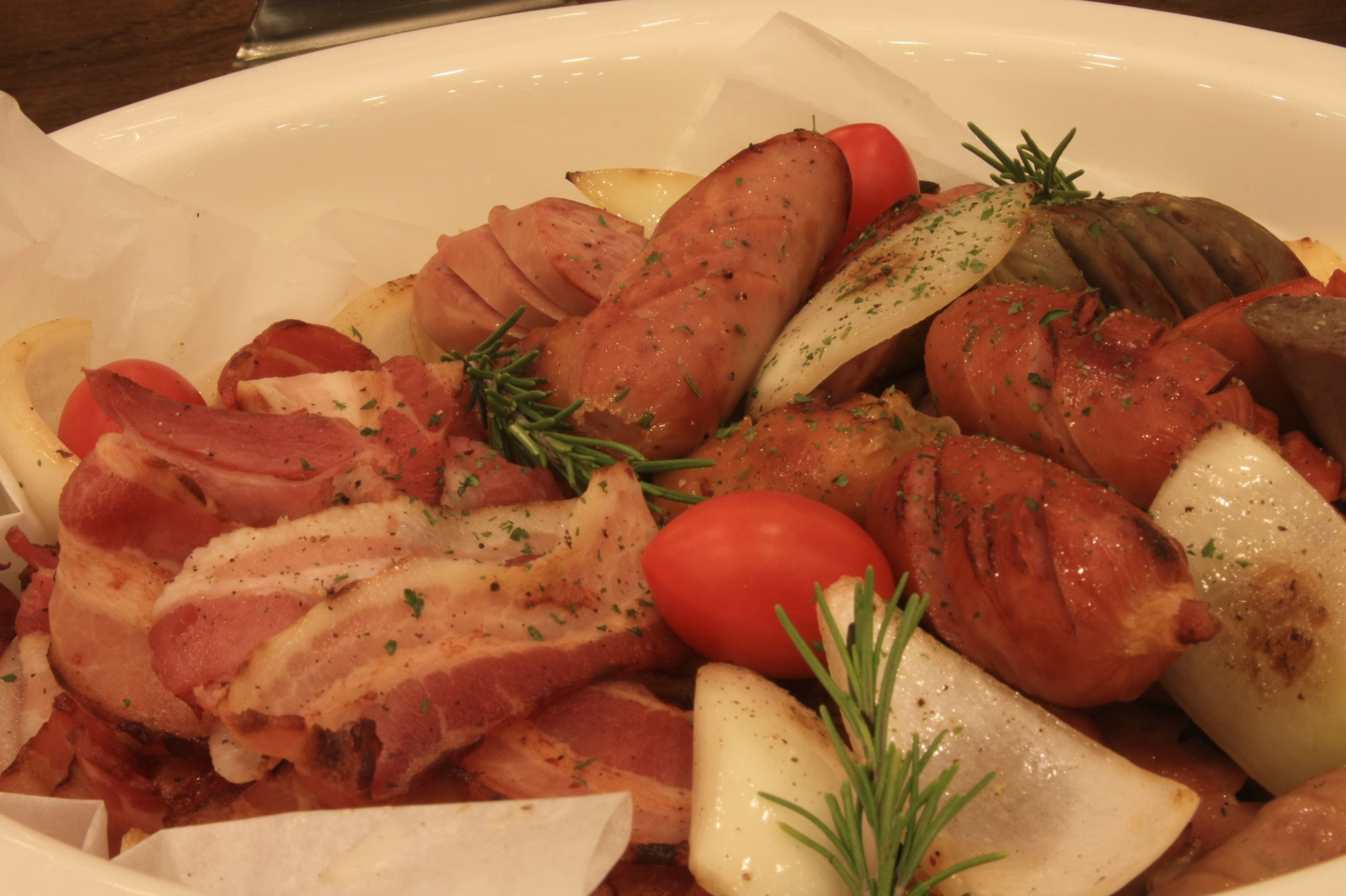 Un plat en céramique blanche contenant des tranches de viande, des pommes de terre et des tomates, le tout garni d’herbes aromatiques.