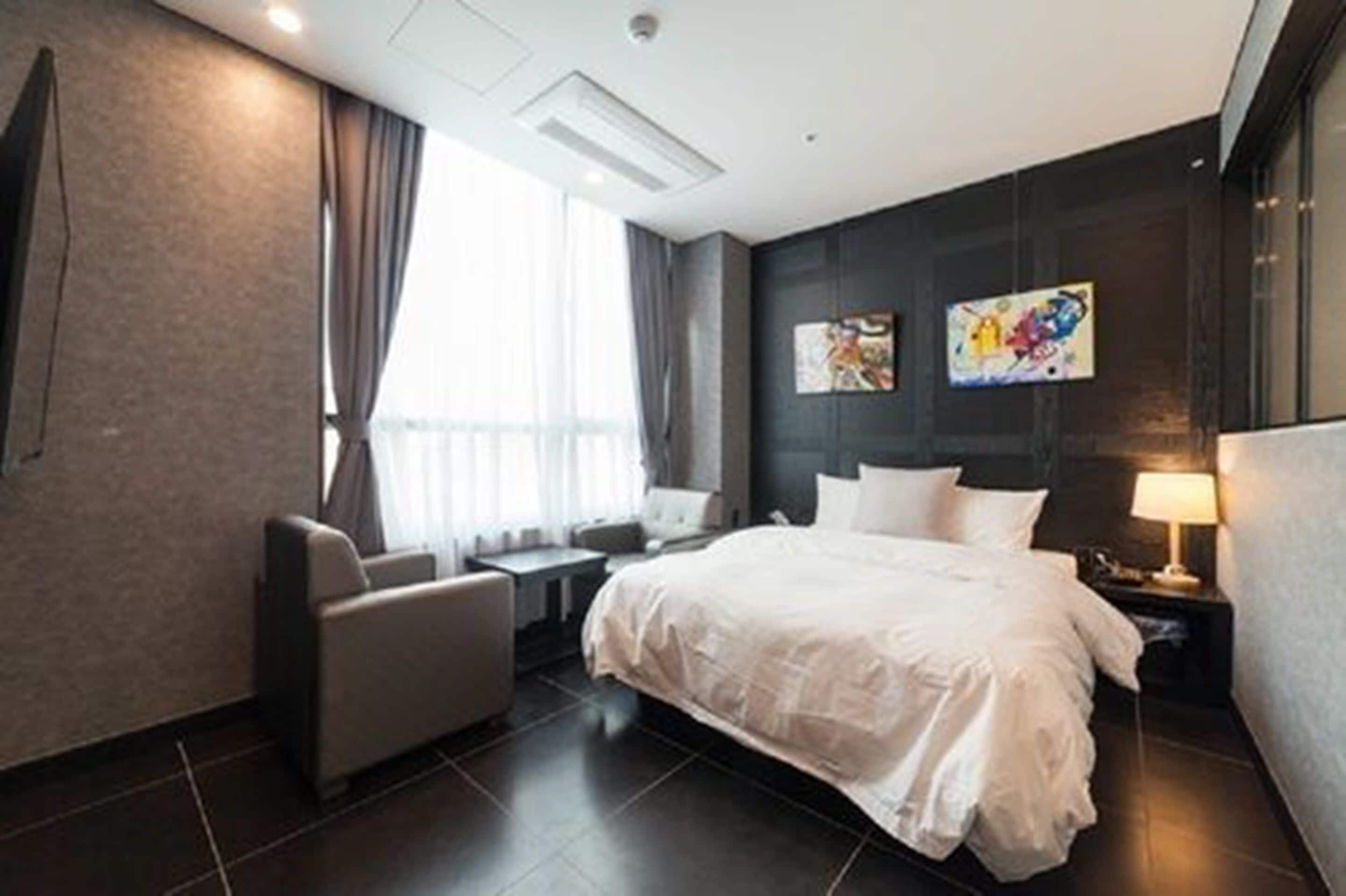 A modern hotel room with a large bed, a desk, a chair, and a window with curtains.