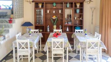 Un comedor con suelo a cuadros, sillas blancas y mesas cubiertas con manteles estampados. En el fondo, un aparador de madera con puertas de cristal y varios objetos decorativos.