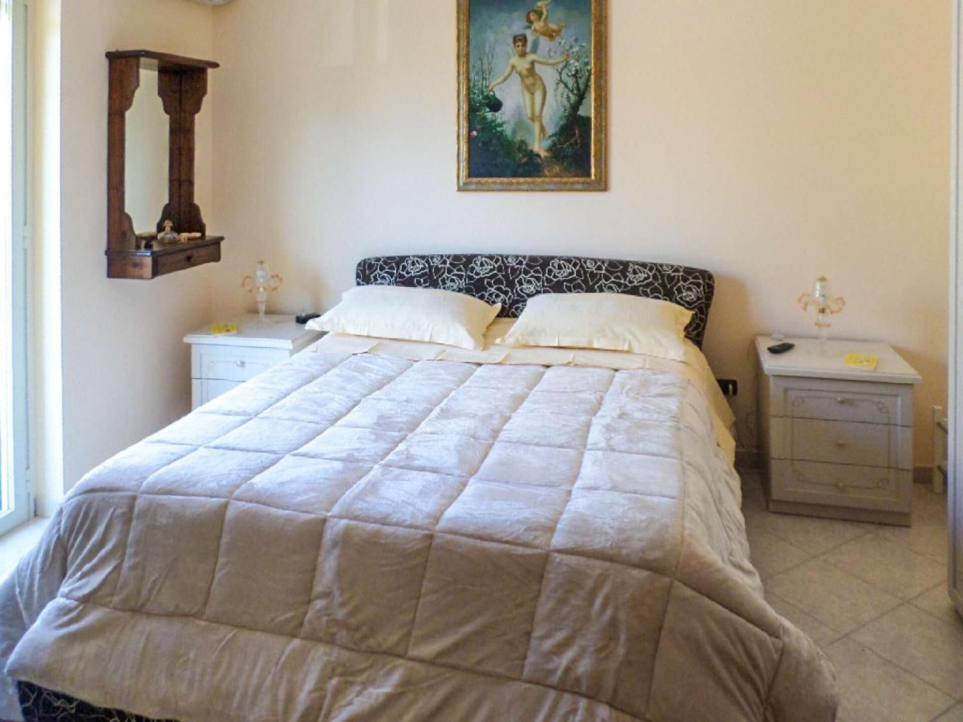 Cama bem arrumada com edredom acolchoado, duas mesas de cabeceira com luminárias, espelho fixado na parede e quadro sobre a cama.