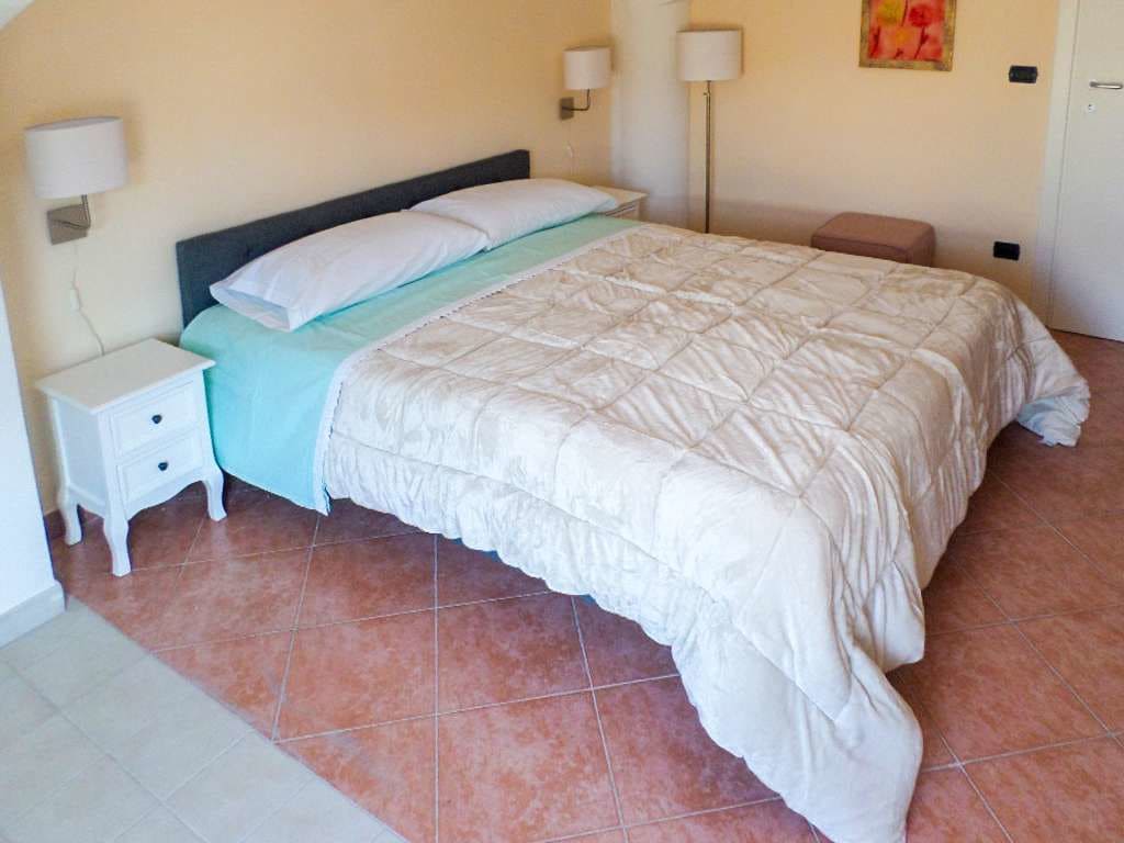Una cama bien hecha con colcha blanca, una mesita de noche pequeña y una lámpara de pared.