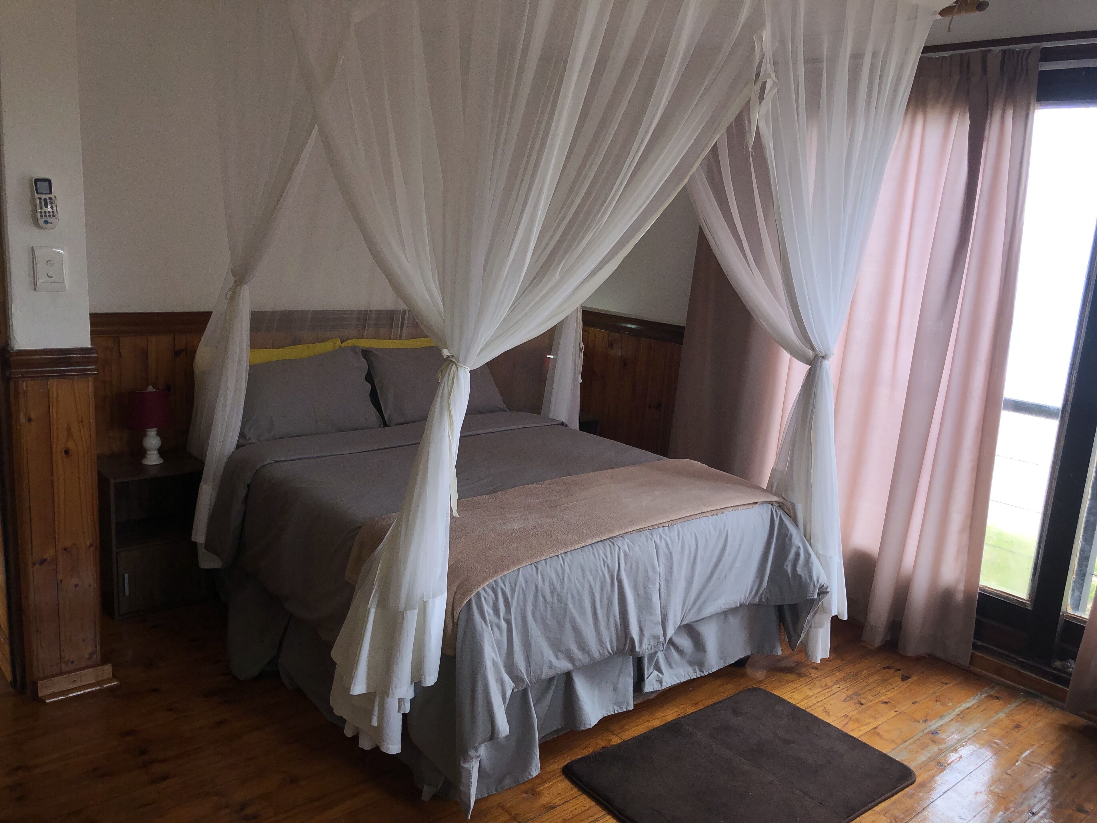 A bedroom with a four-poster bed, wooden flooring, a bedside table with a lamp, and a window with curtains.