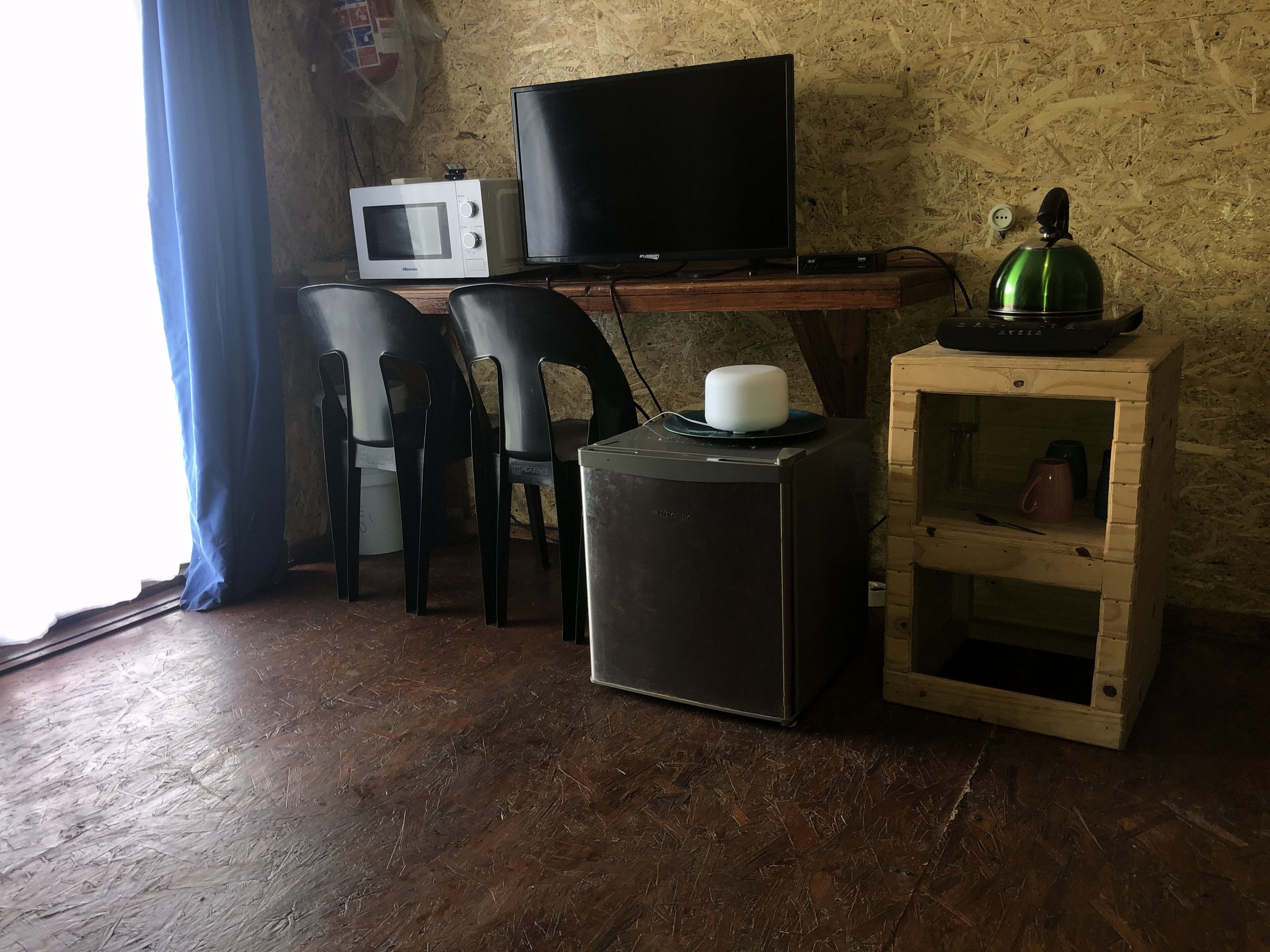 Una habitación con un escritorio de madera, un televisor, un microondas, dos sillas negras, un mueble de madera pequeño y una taza roja.