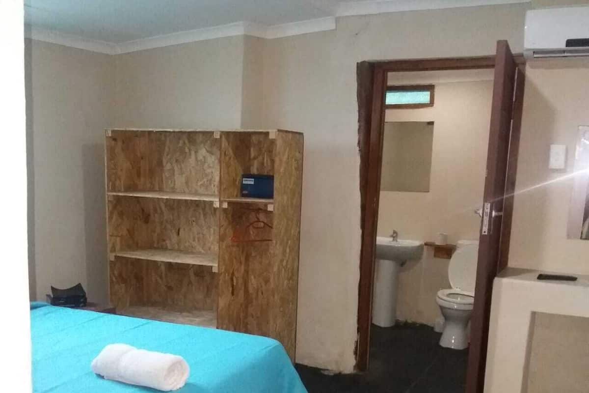 Quarto com cama, guarda-roupa de madeira, banheiro com vaso sanitário e pia, e uma pequena mesa.