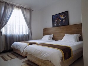 A hotel room with two beds, a wooden headboard, a painting of birds, and a window with curtains. - Remmings Hotel (Kampala)