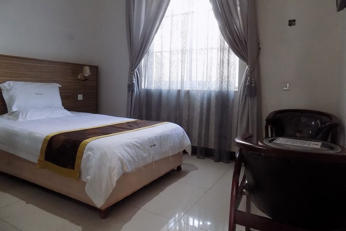 A hotel room with a bed, a chair, a window with curtains, and a wooden headboard.