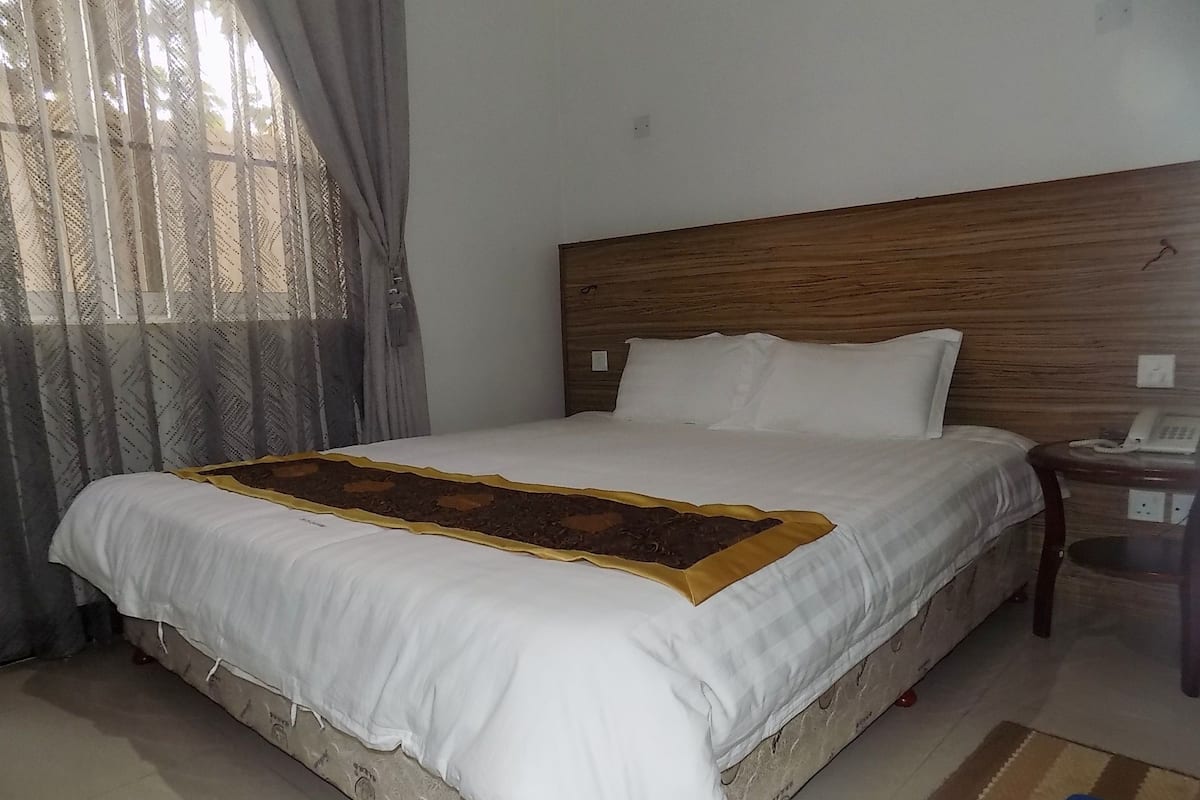 A neatly made bed with a wooden headboard, a bedside table with a telephone, and a window with sheer curtains.