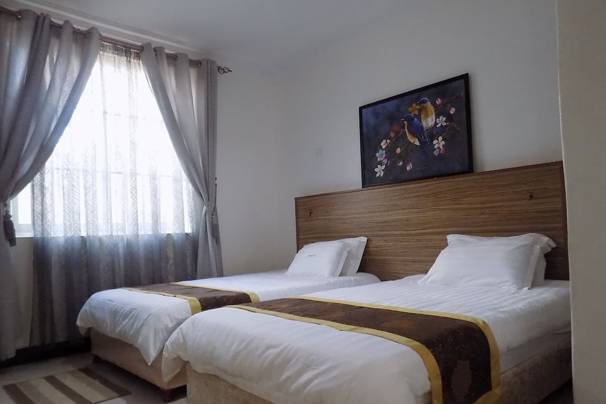 A hotel room with two beds, a wooden headboard, a painting of birds, and a window with curtains.