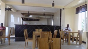 A dining area with wooden tables and chairs, a kitchen in the background, and a large window on the right. - Remmings Hotel (Kampala)