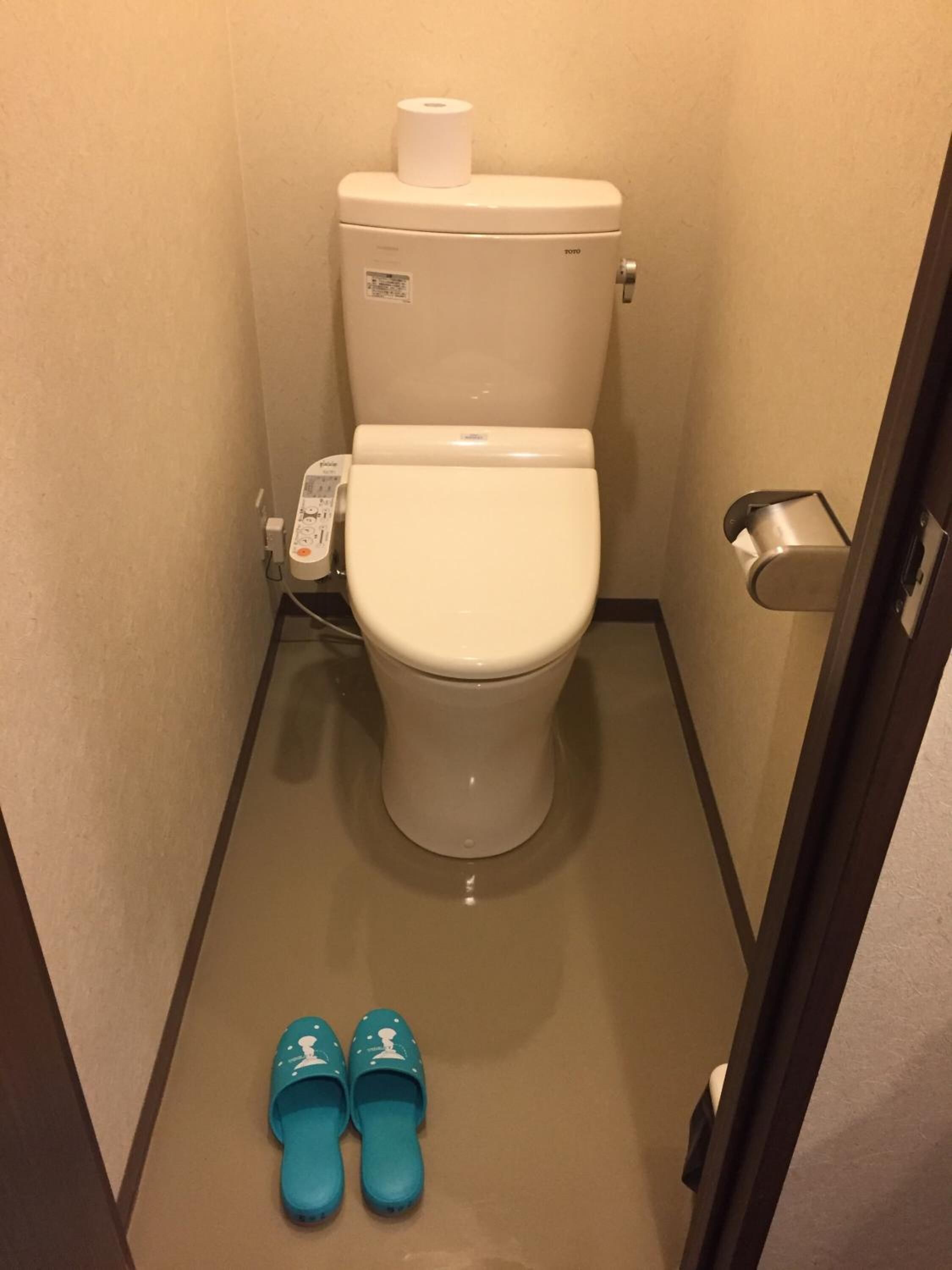 Un baño pequeño con inodoro, dispensador de toallas de papel y un par de pantuflas en el suelo.