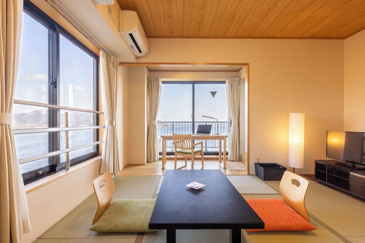Quarto com vista para o mar, uma mesa com cadeiras, uma televisão e uma escrivaninha com um computador.