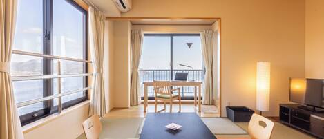 A room with a view of the sea, a table with chairs, a TV, and a desk with a computer.