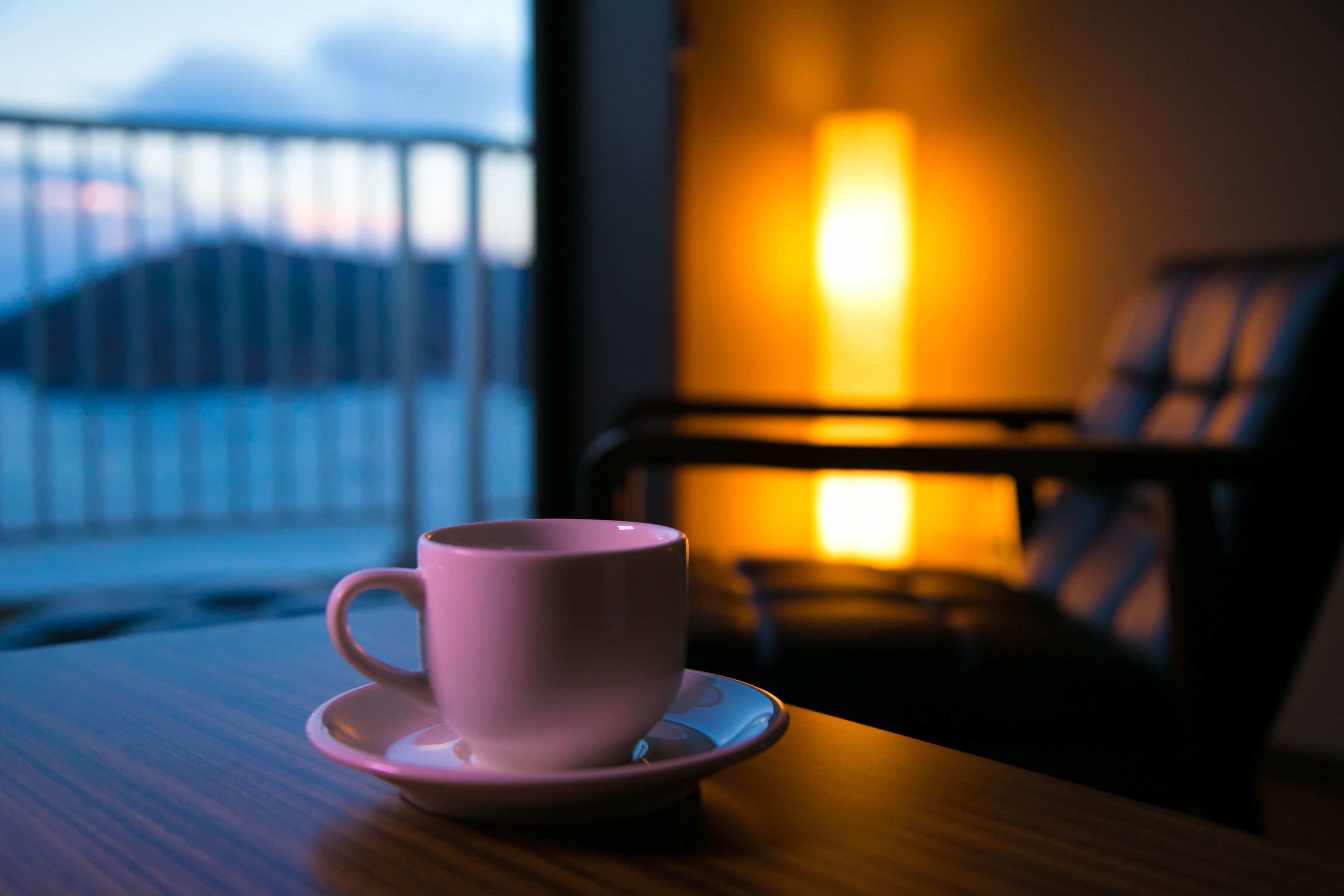 A pink cup on a saucer on a wooden table with a view of a balcony and a lit lamp in the background.