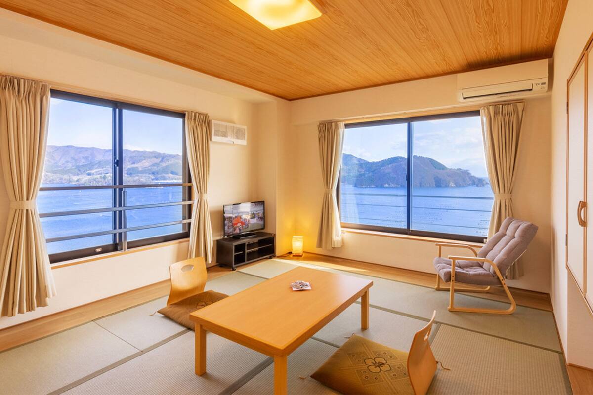 Quarto com teto de madeira, janelas amplas com cortinas, televisão, mesa de madeira, cadeira e esteiras de tatame no chão.