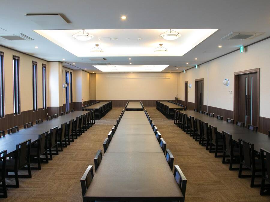 Ein Konferenzraum mit langen Tischen und Stühlen, einer hohen Decke mit eingelassener Beleuchtung und großen Fenstern an einer Seite.