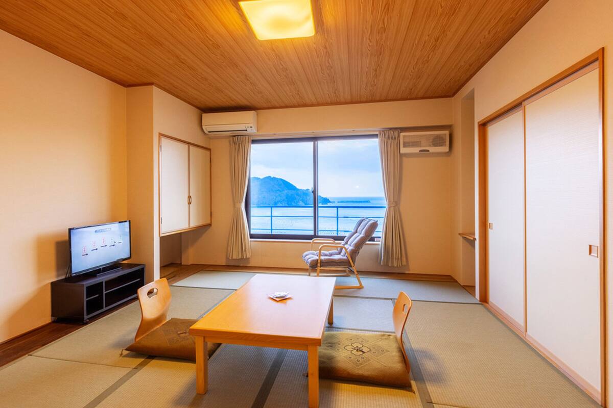 Quarto com teto de madeira, televisão, mesa, cadeiras e vista para o mar, além de uma porta deslizante.