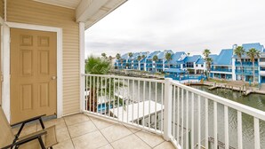 A balcony with a view of a marina, featuring blue buildings and palm trees. - Spacious 2br 2bath condo! On the canal with a boat lift! (Corpus Christi)
