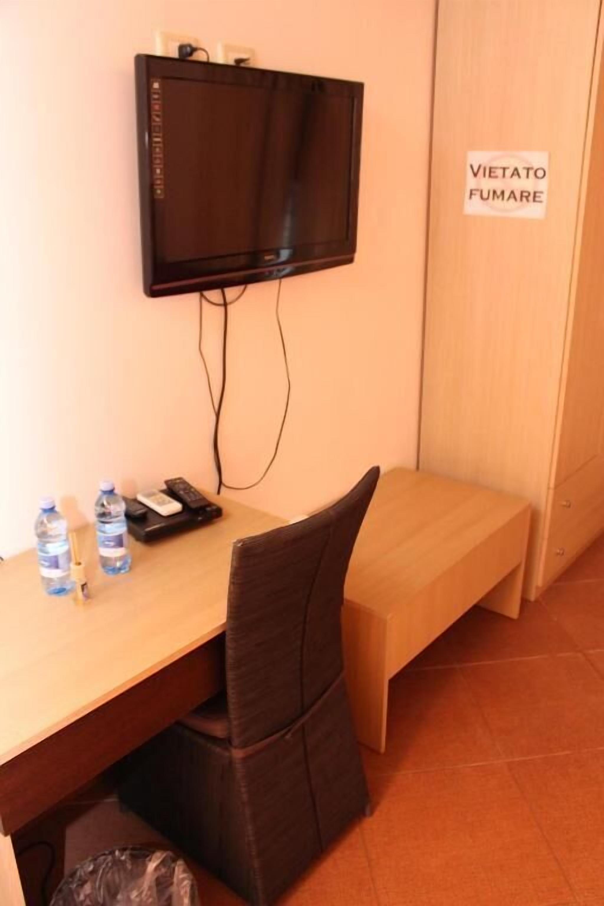 A small room with a desk, a chair, a television mounted on the wall, and a sign reading "Vietato Fumare".