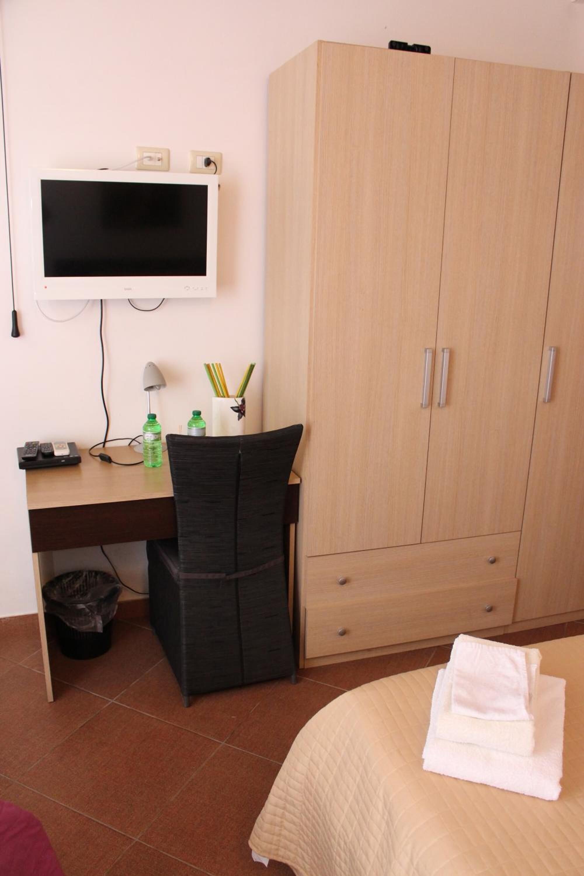 A hotel room with a bed, a desk with a computer monitor, a chair, a wardrobe, and a trash bin.