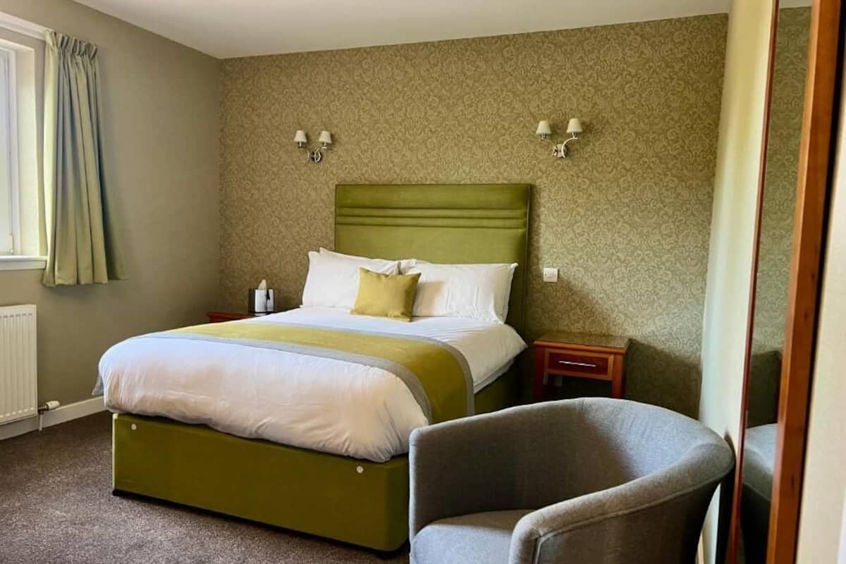 A hotel room with a green upholstered bed, a grey armchair, and patterned wallpaper.