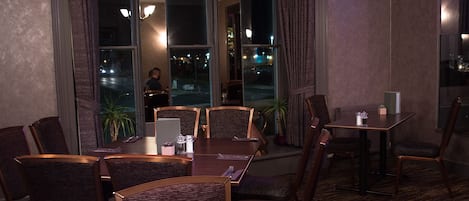 Uma sala de jantar com uma mesa grande e cadeiras, uma mesa menor com uma cadeira e vista para a rua.