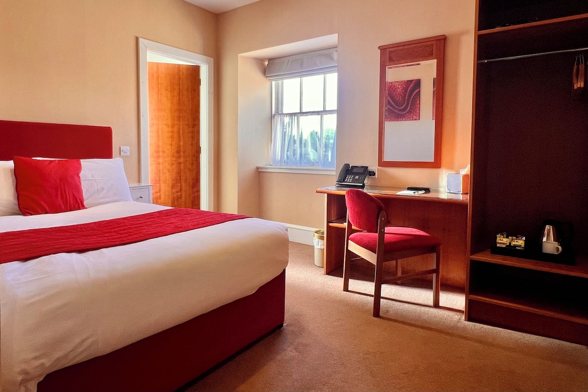 A hotel room with a large bed, a desk with a chair, a wardrobe, and a window with curtains.