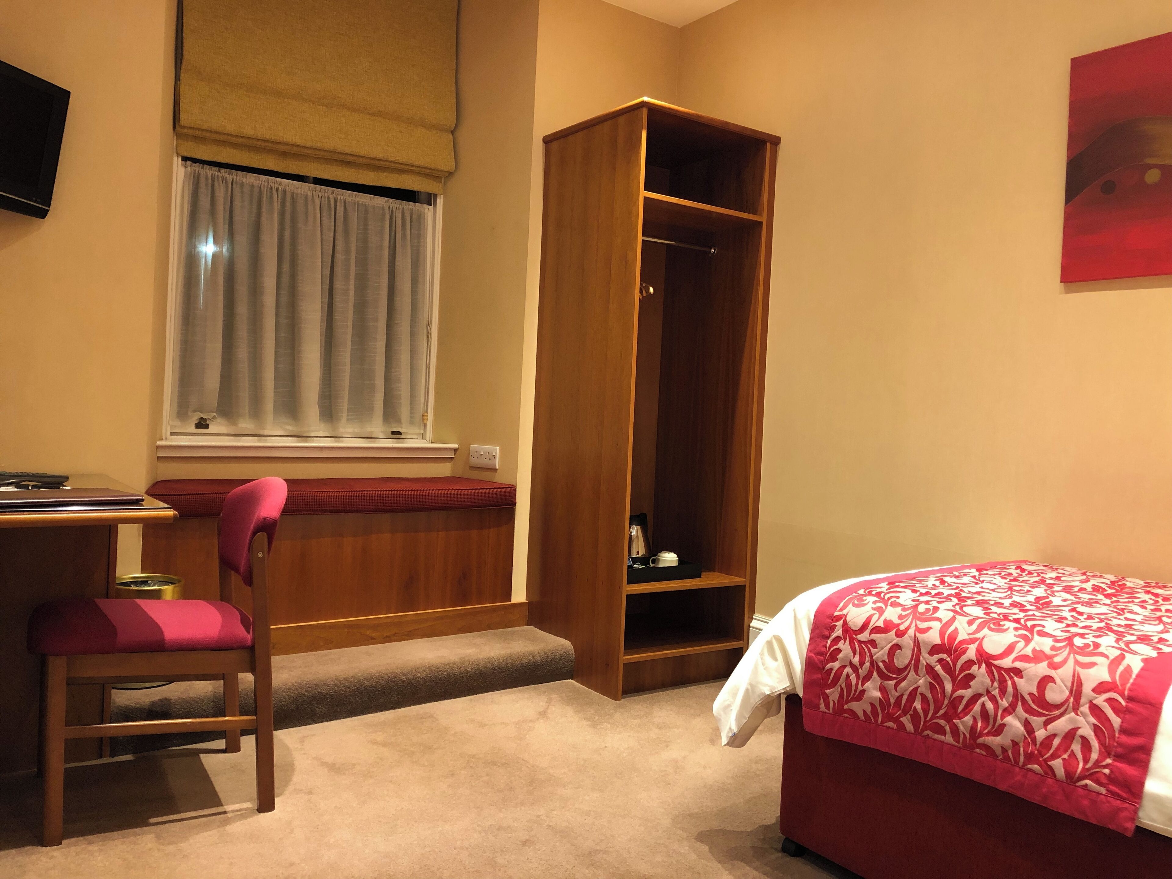 A hotel room with a desk, chair, bed, wardrobe, and a window with curtains.