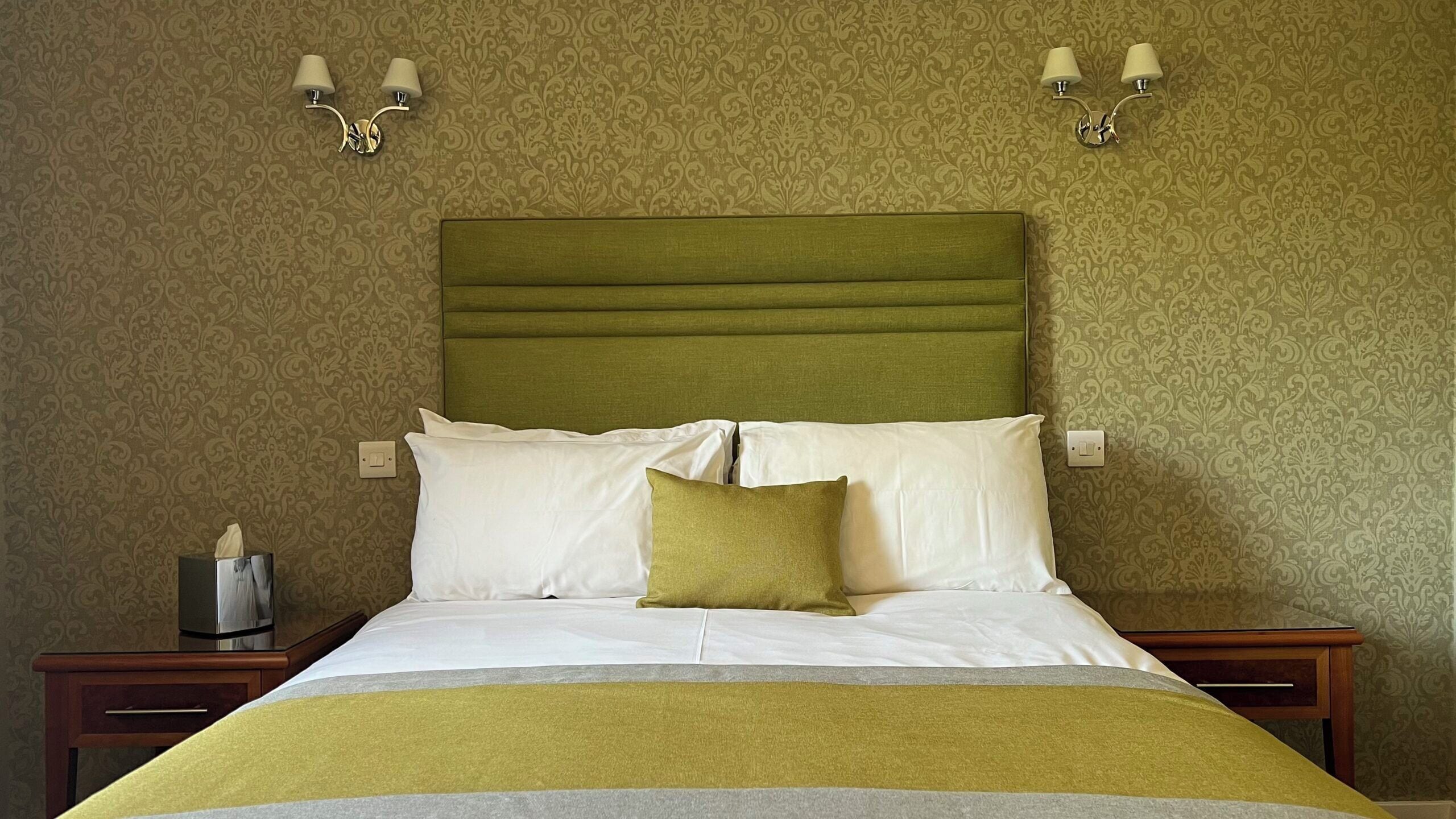 A neatly made bed with a green headboard, white and green pillows, and a yellow blanket. There are two bedside tables with lamps and a tissue box on one.