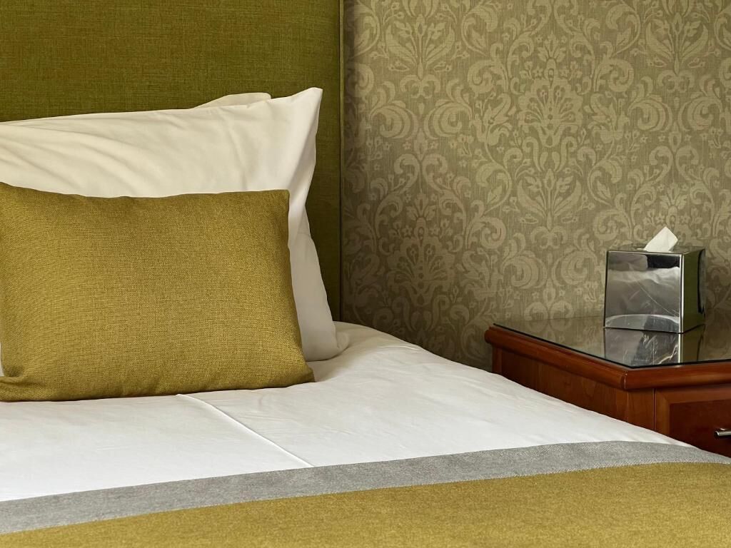 A neatly made bed with white and olive green bedding, a bedside table with a tissue box, and a patterned wallpaper background.