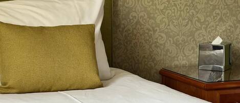 A neatly made bed with white and olive green bedding, a bedside table with a tissue box, and a patterned wallpaper background.
