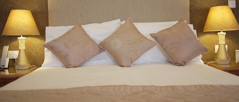 Una cama bien hecha, con tres almohadas beige y un edredón a juego, flanqueada por dos lámparas de noche sobre mesitas de madera.