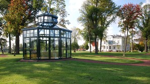 Un belvédère en verre, situé dans un parc, avec un grand bâtiment blanc en arrière-plan.