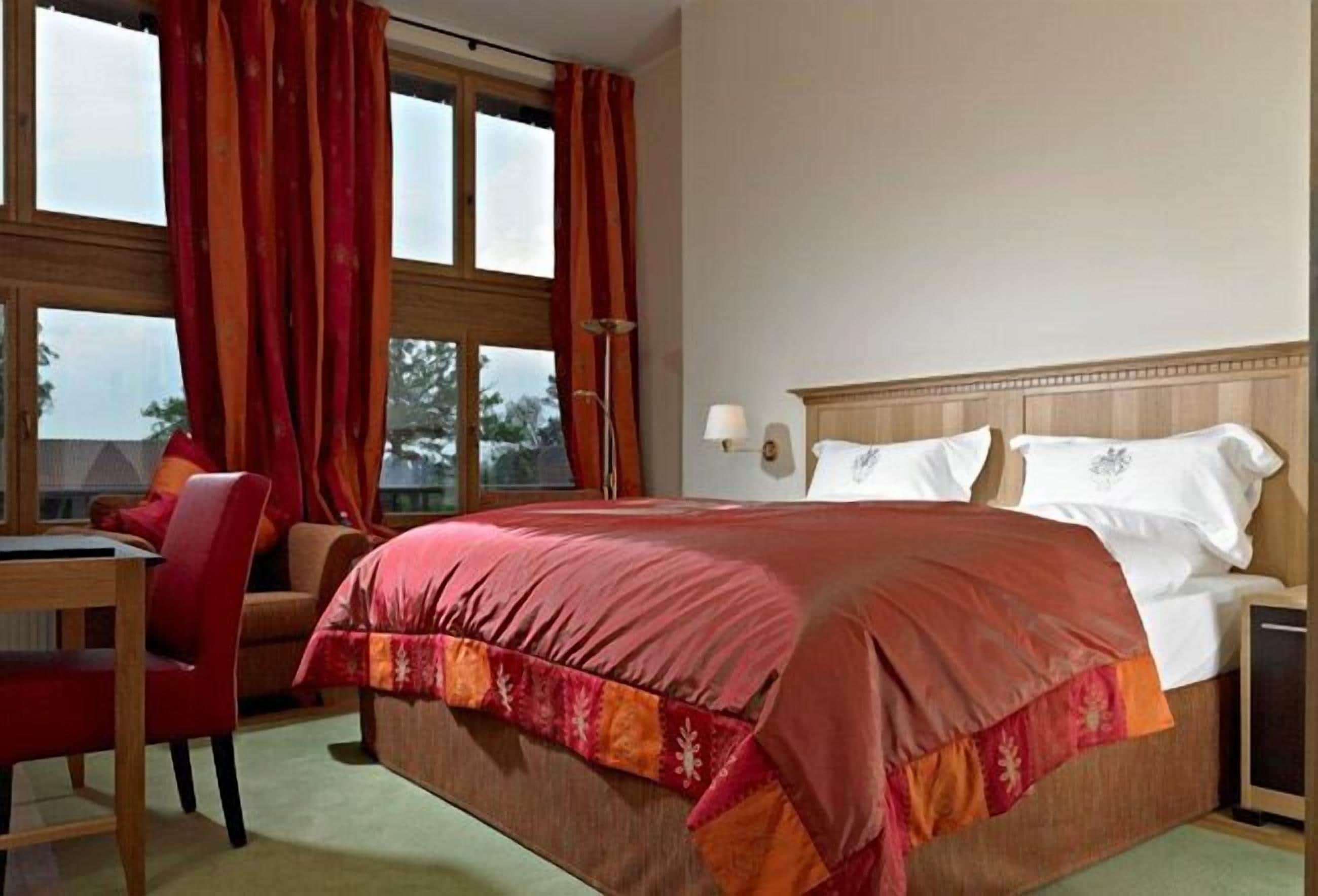 Habitación de hotel con una cama grande, un escritorio y una silla. Hay cortinas en las ventanas, y una lámpara sobre el escritorio.