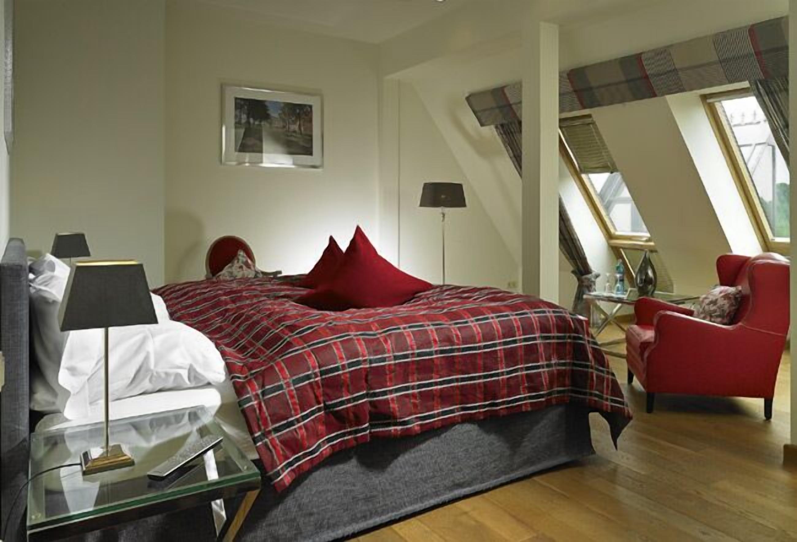 Un dormitorio con una cama, una silla roja, un escritorio con una lámpara y un cuadro en la pared.
