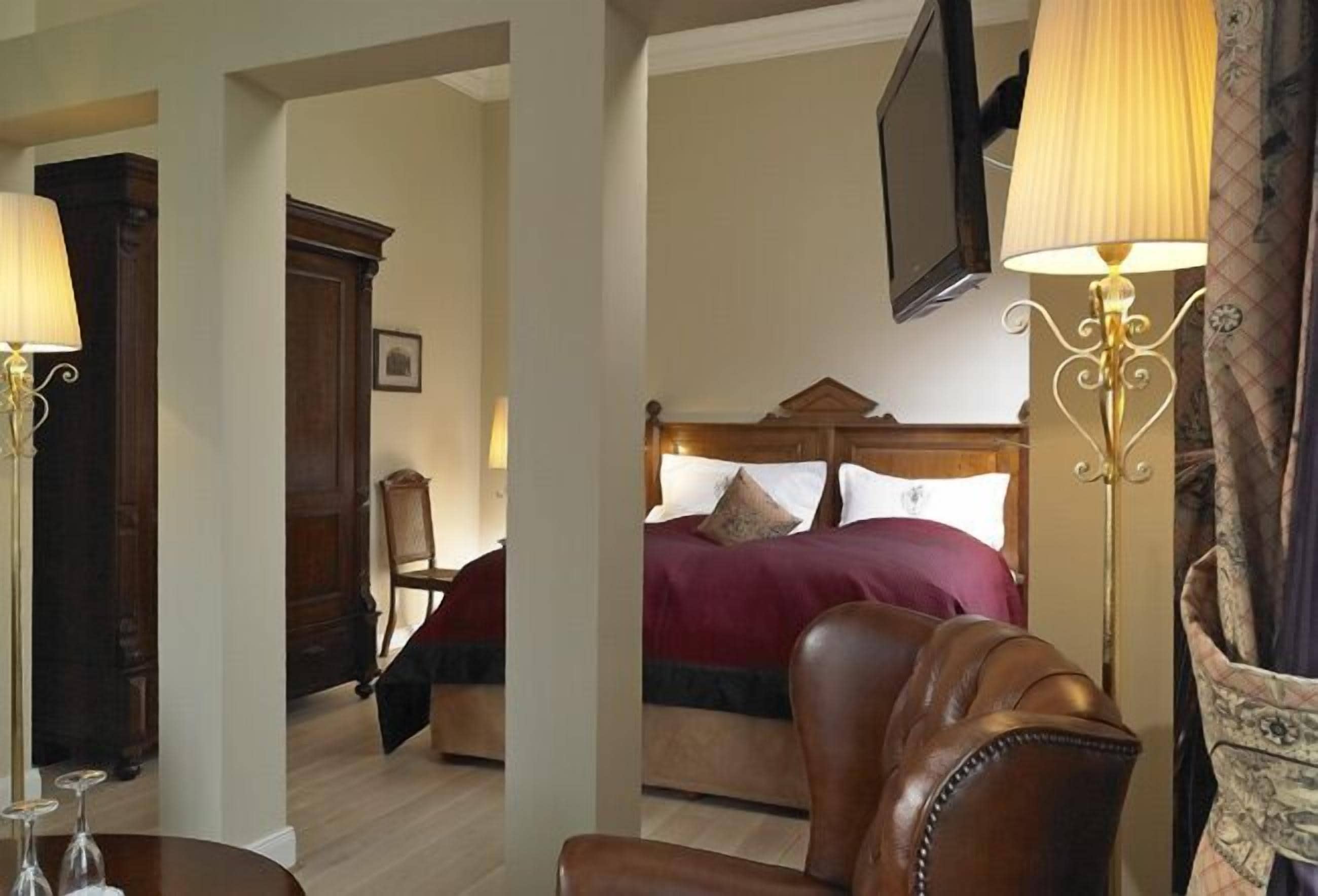 A hotel room with a wooden bed, a leather armchair, a television mounted on the wall, and a lamp.