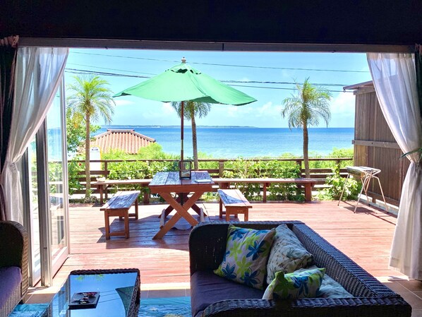 A patio with a wicker sofa, a glass table, and a wooden bench. There is a green umbrella providing shade. The patio overlooks a body of water and greenery. - Suite Terrace Ishigakijima (Ishigaki)