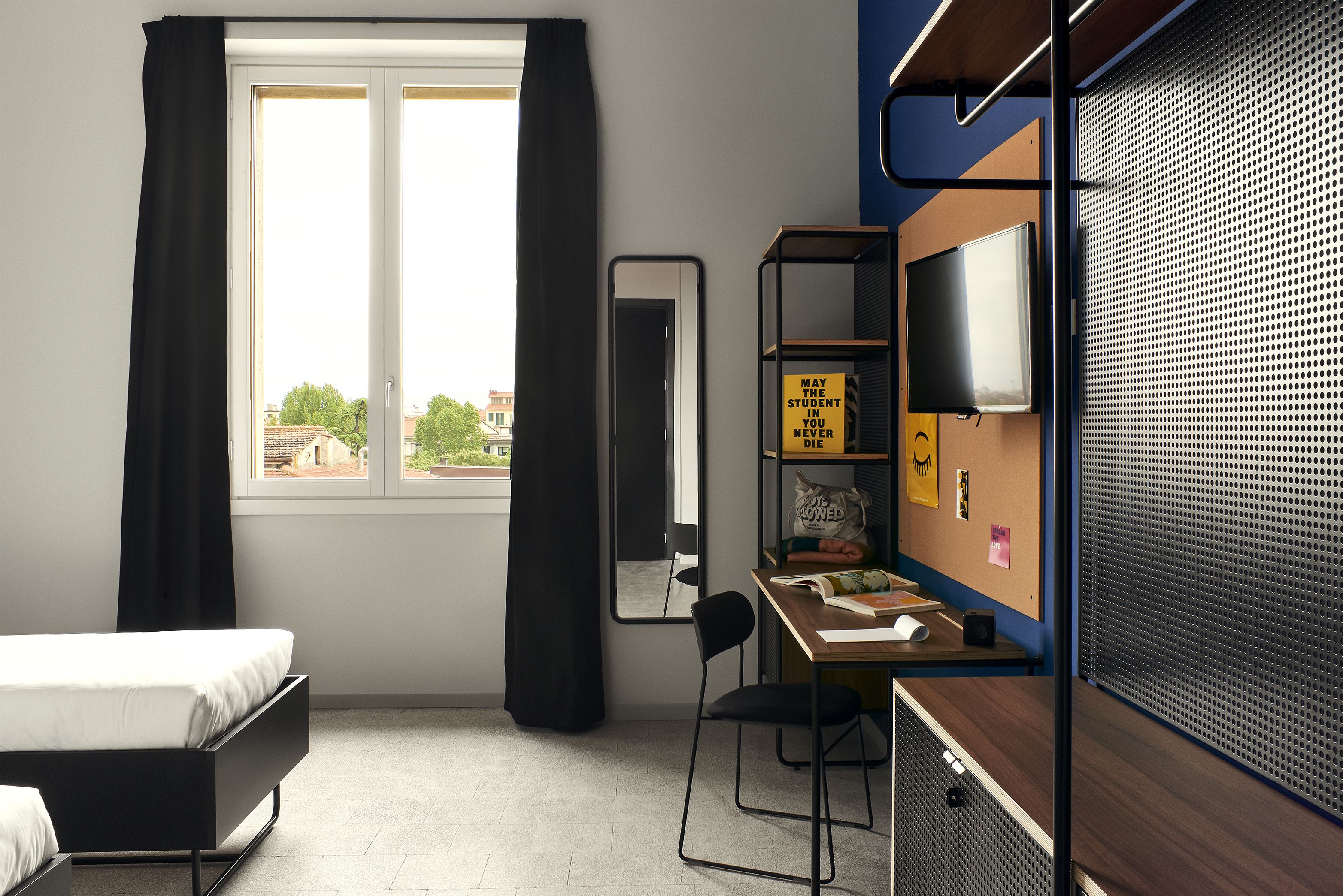 A modern dormitory room with a bed, desk, chair, and a large window with curtains.