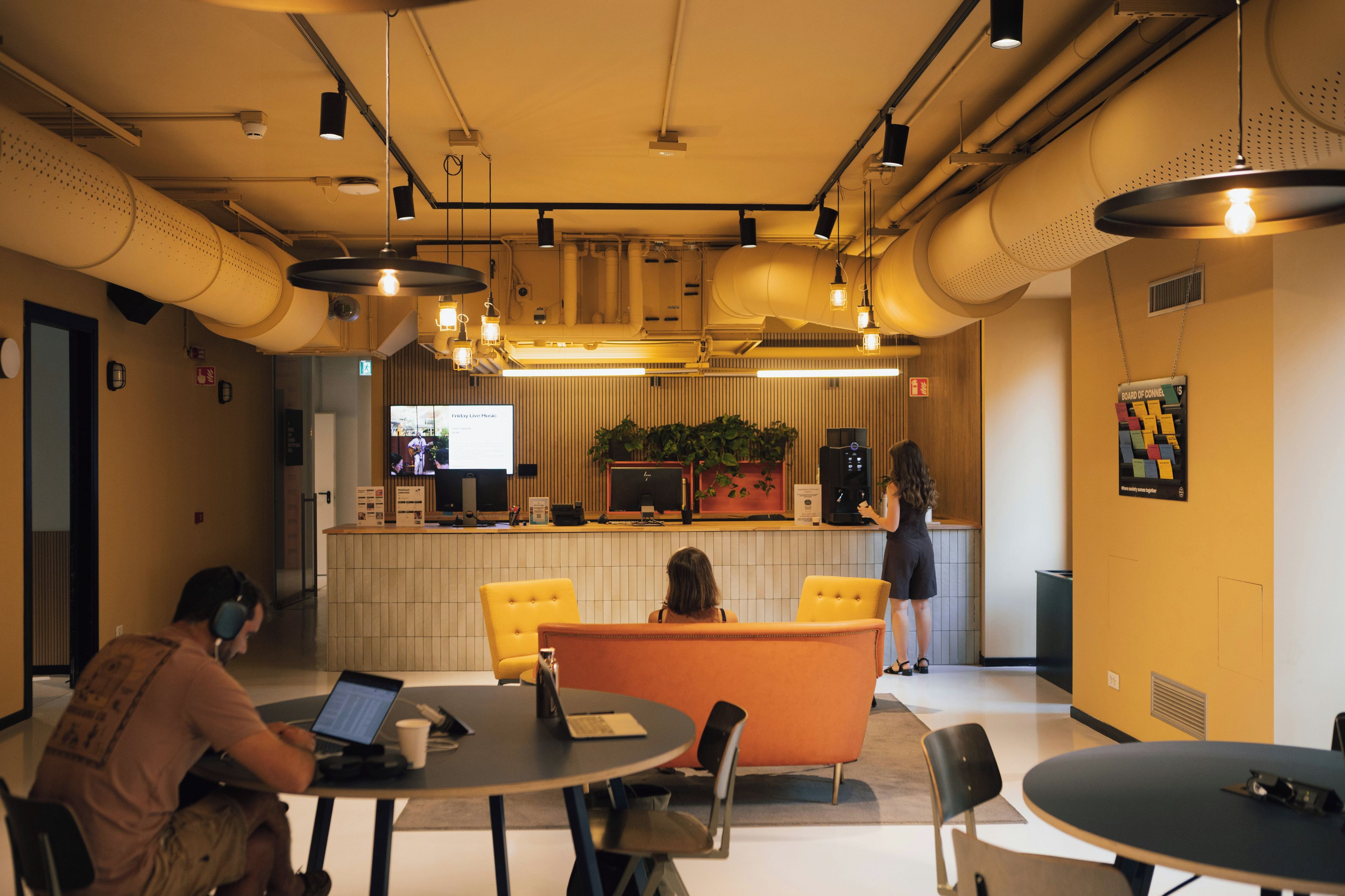 A modern interior with round tables, chairs, a reception desk, and a television screen displaying 'Friday Live Music'.